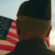 An older soldier standing with his back to the camera watching an American flag wave.